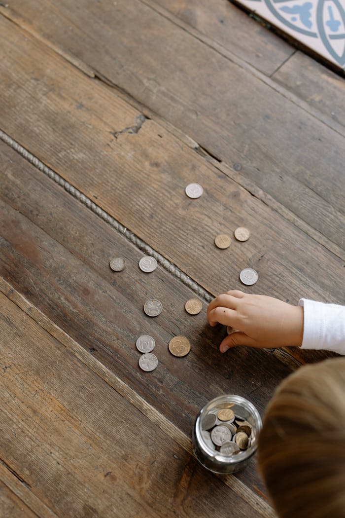 Overhead view of child's hand gathering coins into a glass jar on wooden flooring.